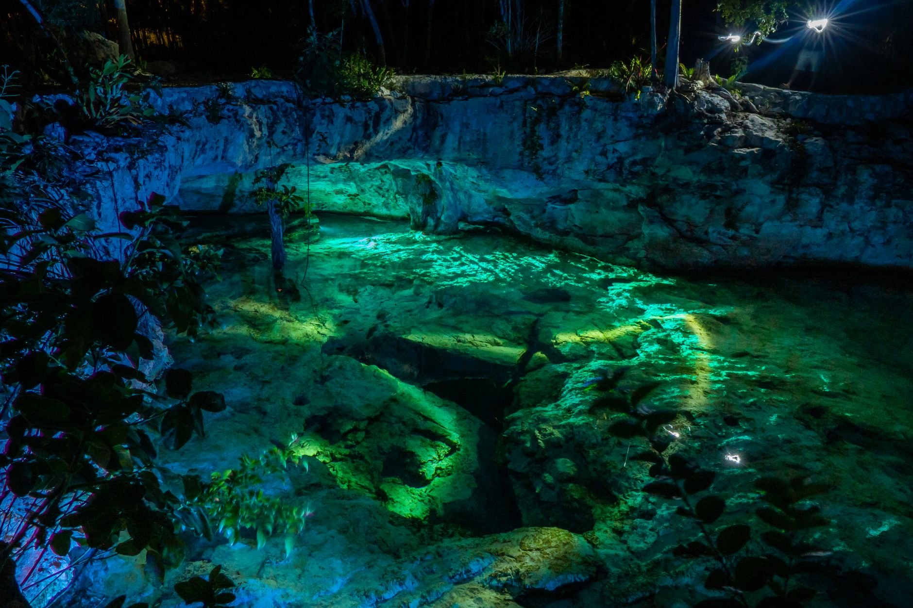 Illuminated cenote in Tulum, Mexico at night — turquoise water glowing beneath rocky walls